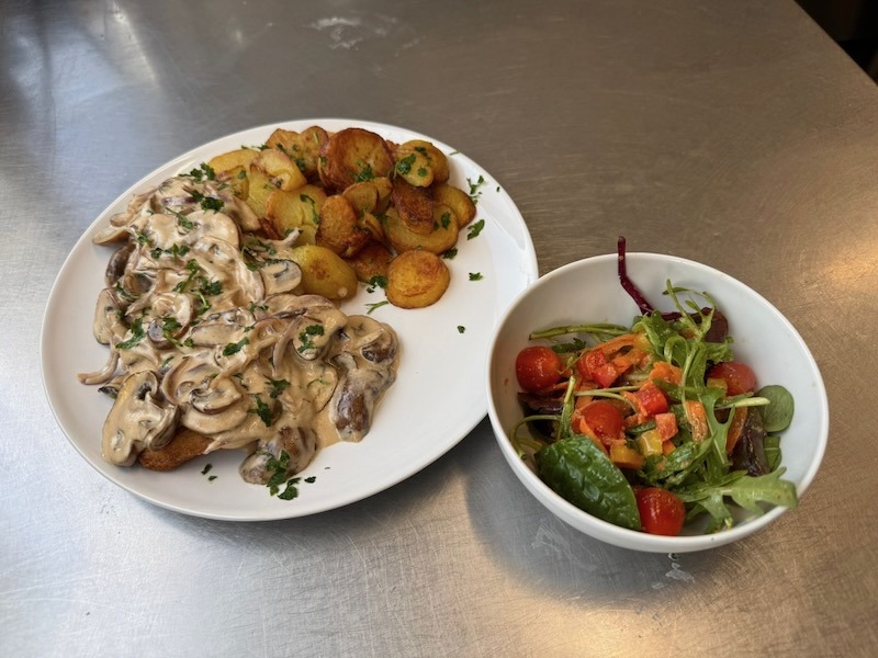 Paniertes Schnitzel mit cremiger Pilz-Rahmsauce und frischer Petersilie auf einem weißen Teller, dazu Bratkartoffeln. Als Beilage eine Schüssel Blattsalat mit Kirschtomaten und Paprika.