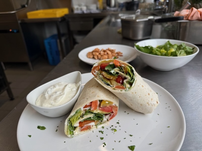 Aufgeschnittener Gemüse-Wrap aus Weizenfladen mit buntem Pfannengemüse – Paprika, Zucchini, Tomaten und Salat – auf einem weißen Teller, dazu ein Schälchen Joghurt-Dip mit frischen Kräutern.