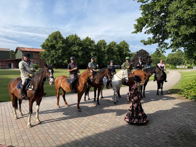 Sechs Reiter der Herrenquadrille von 1979 zu Pferd am Hofrondell von Gut Tangstedt. Eine Dame im geblümten spanischen Kleid empfängt die Gruppe mit einem Glas Sherry. Im Hintergrund die gepflegte Gutsanlage mit altem Baumbestand bei Abendlicht.
