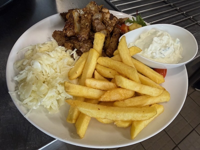 Gyros-Pfanne mit gewürztem Schweinefleisch und Paprika auf einem weißen Teller, serviert mit Pommes frites, Krautsalat und einem Schälchen Tzatziki.