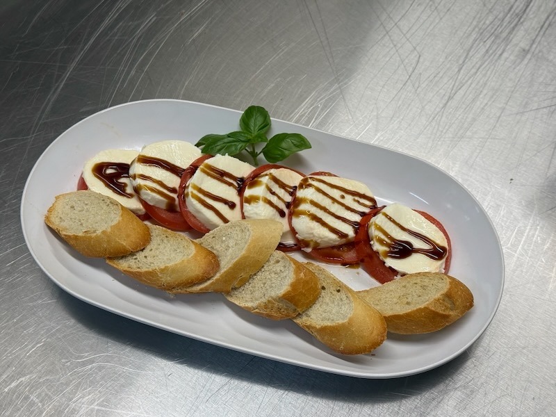 Klassischer Caprese auf einem ovalen wei&szlig;en Teller: abwechselnde Scheiben von Tomate und Mozzarella mit Balsamico-Reduktion und frischem Basilikum, dazu ger&ouml;stete Baguettescheiben.