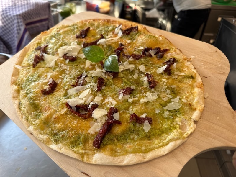 Knusprig gebackener Flammkuchen mit Basilikumpesto und getrockneten Tomaten auf einem Holzbrett, garniert mit frischen Basilikumblättern.