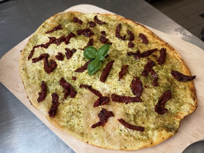 Knusprig gebackener Flammkuchen mit Basilikumpesto und getrockneten Tomaten auf einem Holzbrett, garniert mit frischen Basilikumbl&auml;ttern.