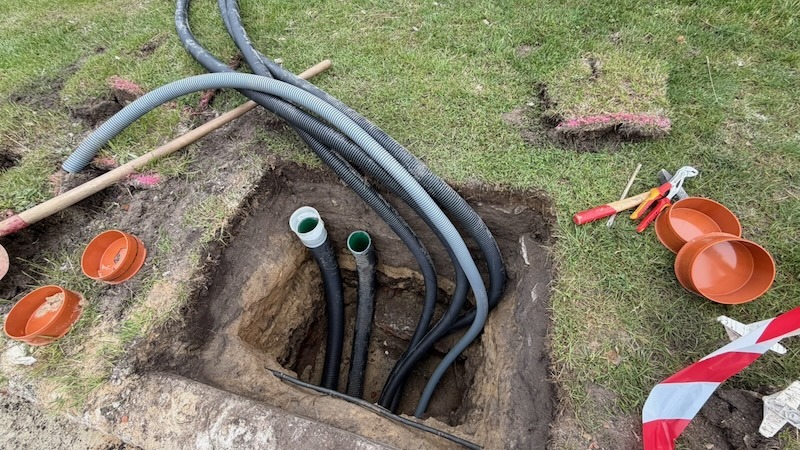 Offene Baugrube im Rasen mit mehreren eingelegten Leerrohren und Schutzrohren für die Kabelverlegung. Neben der Grube liegen Kanaldeckel, Werkzeug und rot-weißes Absperrband.