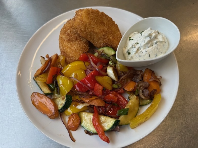 Zwei knusprig panierte Feta-Scheiben auf einer bunten mediterranen Gemüsepfanne mit Paprika, Zucchini, Karotten und Frühlingszwiebeln, dazu ein Schälchen hausgemachter Kräuterquark.