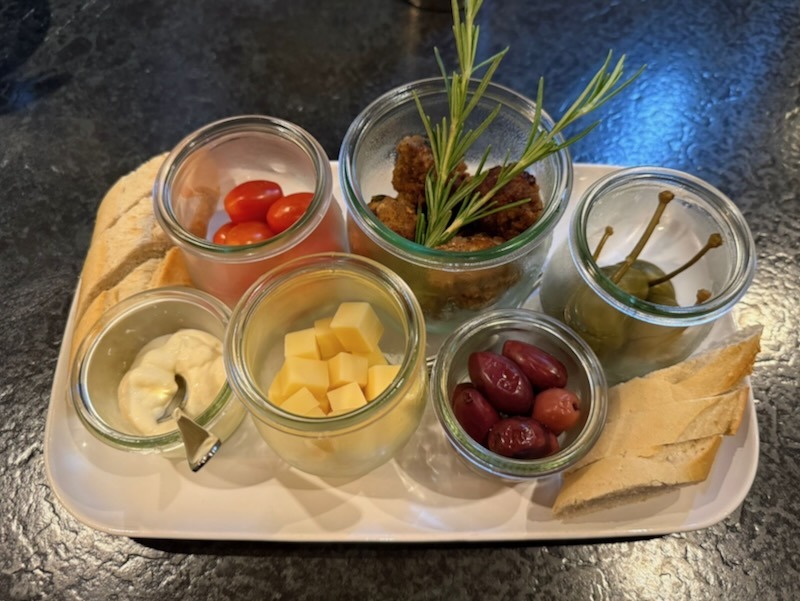 Antipasti-Platte mit mehreren Schälchen: schwarze Oliven, gewürfelter Käse, gebratene Fleischbällchen, Kirschtomaten, Kapern sowie ein cremiger Dip, dazu Weißbrotscheiben.