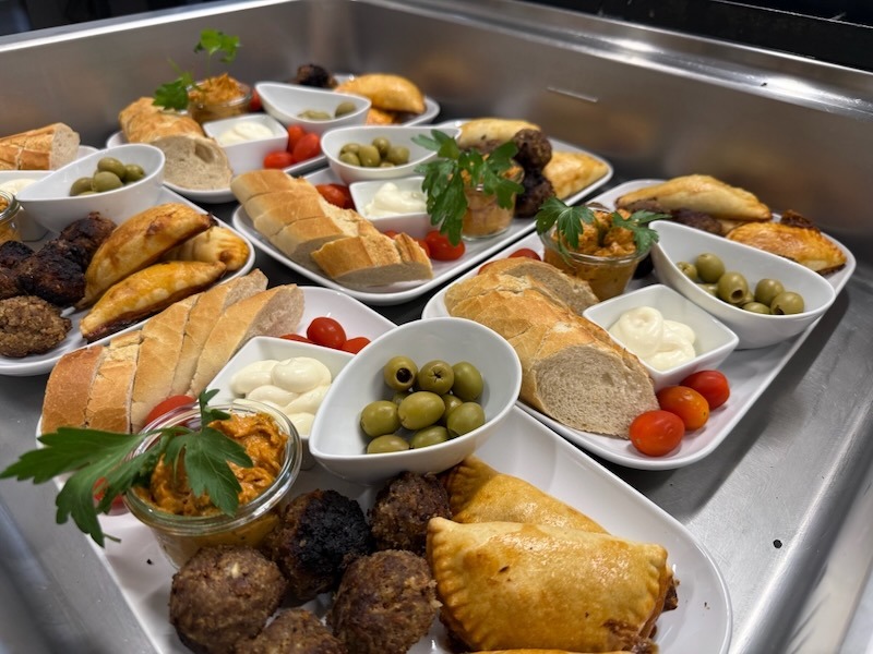 Tapas-Variationen auf einem Tablett: Empanadas, Hackbällchen, Weißbrot, Aioli, grüne Oliven, Cocktailtomaten und Paprika-Dip in einem kleinen Glas.