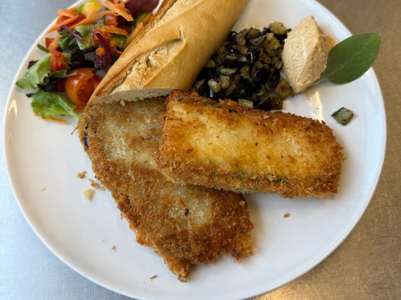Zwei knusprig panierte Auberginen-Zucchini-Schnitzel auf einem wei&szlig;en Teller, serviert mit geschmorter Aubergine, Hummus, Baguette, Blattsalat mit Kirschtomaten und Karotten sowie einem frischen Salbeiblatt.