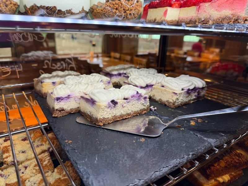 Blaubeer-Cheesecake-Schnitten mit M&uuml;rbeteigboden und Baiserrosetten auf einer Schieferplatte in der Kuchenvitrine der Hofk&uuml;che.