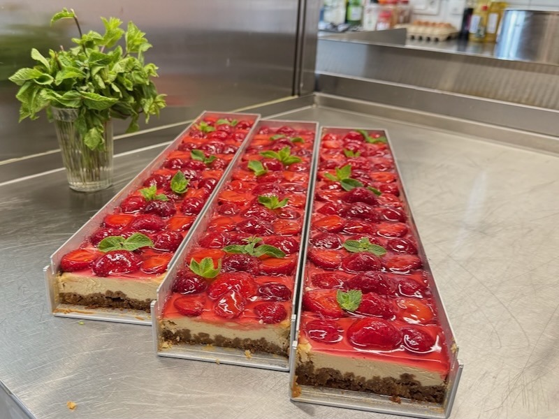 Drei frisch zubereitete Erdbeer-Cheesecake-Riegel mit gl&auml;nzendem Fruchtguss und frischer Minze auf der Edelstahlarbeitsplatte der Hofk&uuml;che.