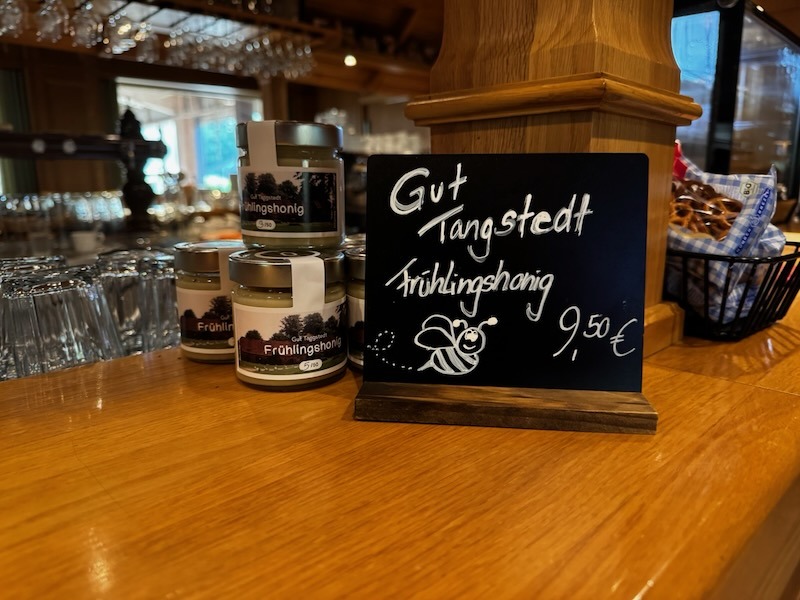 Gläser des Gut Tangstedt Frühlingshonigs auf der Theke der Hofküche, daneben eine Kreidetafel mit der Aufschrift "Gut Tangstedt Frühlingshonig 9,50 €" und einer gezeichneten Biene.