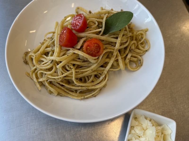 Linguine mit Salbeibutter auf einem wei&szlig;en Teller, garniert mit halbierter Kirschtomate und einem frischen Salbeiblatt, dazu ein Sch&auml;lchen gehobelter Parmesan.