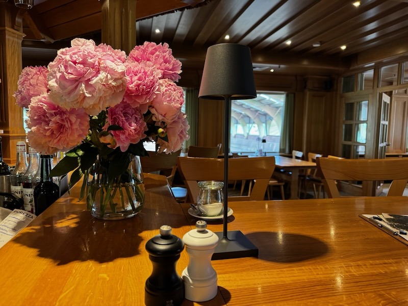 Großer Strauß rosa Pfingstrosen in einer Glasvase auf dem Tresen der Hofküche, daneben eine schwarze Tischlampe sowie Salz- und Pfeffermühle. Im Hintergrund der holzvertäfelte Gastraum mit gedeckten Tischen.