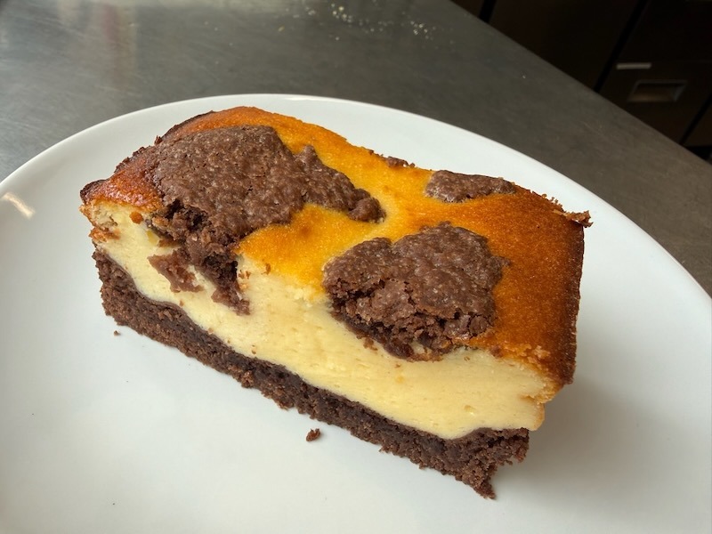 Aufgeschnittenes St&uuml;ck Zupfkuchen mit dunklem Schokoboden und marmorierter K&auml;semasse auf einem wei&szlig;en Teller.