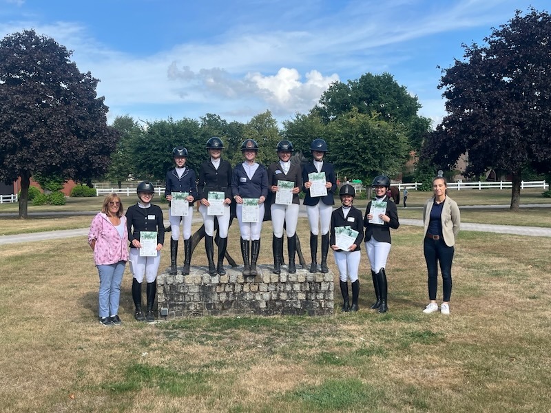 Gruppenbild auf dem Hof-Rondell von Gut Tangstedt: mehrere Reiterinnen in schwarzer Turnierkleidung mit Reithelm stehen auf einem Steinsockel und davor, an den Seiten zwei Frauen in Alltagskleidung. Alle halten ihre Urkunden in den Händen, im Hintergrund Bäume, weiße Koppelzäune und blauer Himmel.