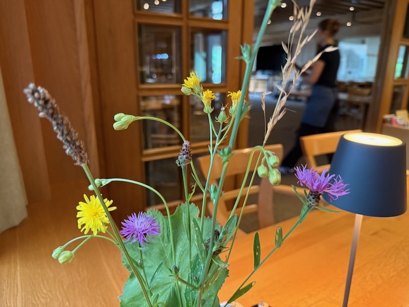 Sommerlicher Wildblumenstrauß mit gelben und lila Blüten sowie Gräsern als Tischdekoration in der Hofküche Gut Tangstedt, im Hintergrund die Servicetheke und eine schwarze Tischlampe.