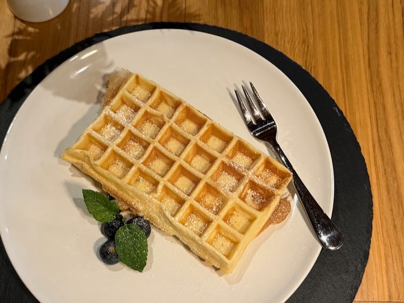 Frisch gebackene belgische Waffel mit Puderzucker, Blaubeeren und Minze auf einem weißen Teller mit Schieferunterlage in der Hofküche Gut Tangstedt.