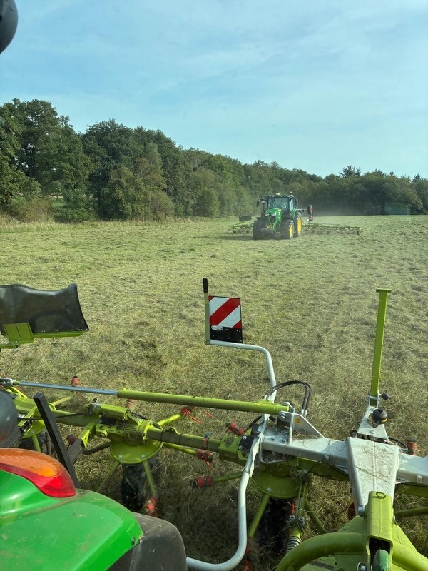 Blick aus der Traktorkabine auf die Heuernte des zweiten Schnitts 2026 auf Gut Tangstedt: Ein John Deere Traktor wendet das gemähte Gras mit einem Kreiselzettwender, im Hintergrund Waldrand bei sonnigem Wetter.