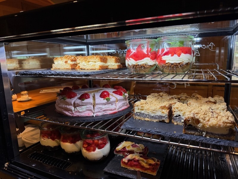 Vollbestückte Kuchenvitrine der Hofküche Gut Tangstedt mit Erdbeertorte, Mohn-Käsekuchen, Streuselkuchen, Desserts im Weckglas und Windbeuteln.