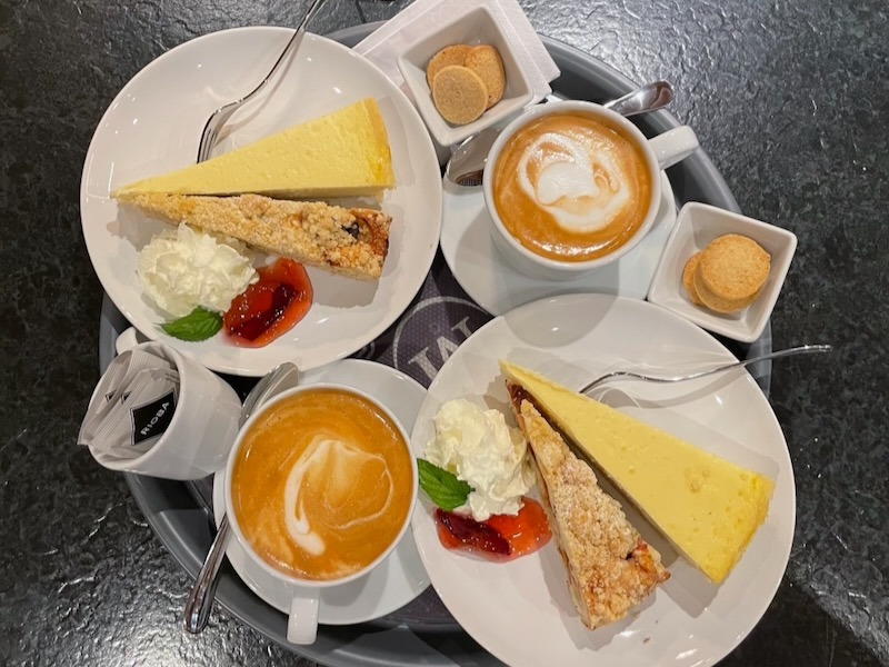 Tablett mit zwei Tassen Cappuccino und zwei Tellern mit je einer Scheibe Käsekuchen und Streuselkuchen mit Schlagsahne und Erdbeersauce – Kaffee und Kuchen in der Hofküche Gut Tangstedt.
