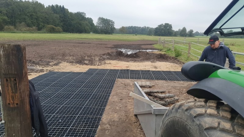 Neu verlegte schwarze Paddockmatten am Eingang der Wallachweide auf Gut Tangstedt, daneben ein grüner Traktor mit weiteren Matten in der Schaufel, im Hintergrund Weidezaun und Wiesen unter bewölktem Himmel