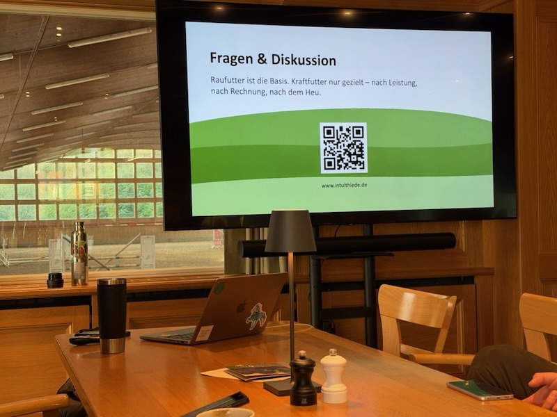 Bildschirm mit der Abschlussfolie „Fragen & Diskussion“ beim Fachvortrag über Kraftfutter auf Gut Tangstedt, davor ein Holztisch mit Laptop, im Hintergrund der Blick durch die Fensterfront in die Reithalle.