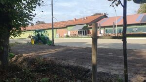 Grüner Traktor auf dem Außenplatz von Gut Tangstedt, dahinter die Hofküche mit rotem Dach, Solarmodulen und einer Terrasse mit Tischen und Sonnenschirmen; im Vordergrund ein Holzpfosten und ein junger Baum am Reitweg